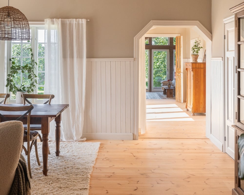 Ett rum med köksbord och stolar, vit bröstpanel och trägolv, samt en öppning till ett annat rum med mycket ljusinsläpp