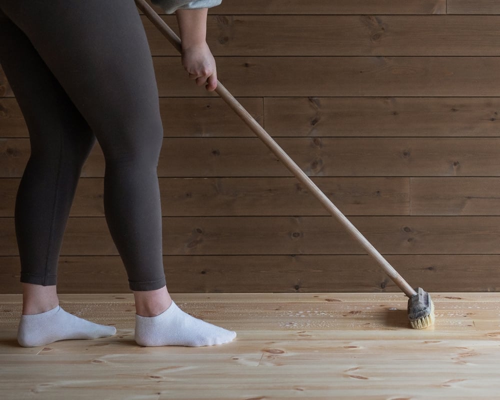 Benen av en kvinna syns medans hon rengör ett trägolv med en borste