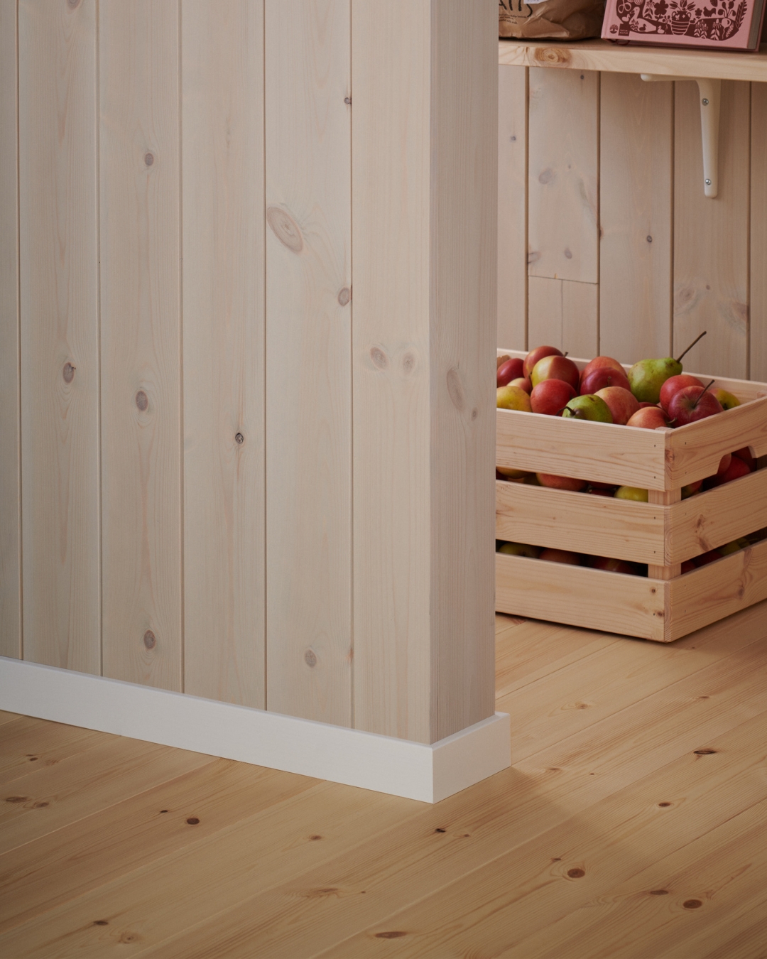 Vägg med panel, trägolv och vita lister, samt en trälåda med äpplen