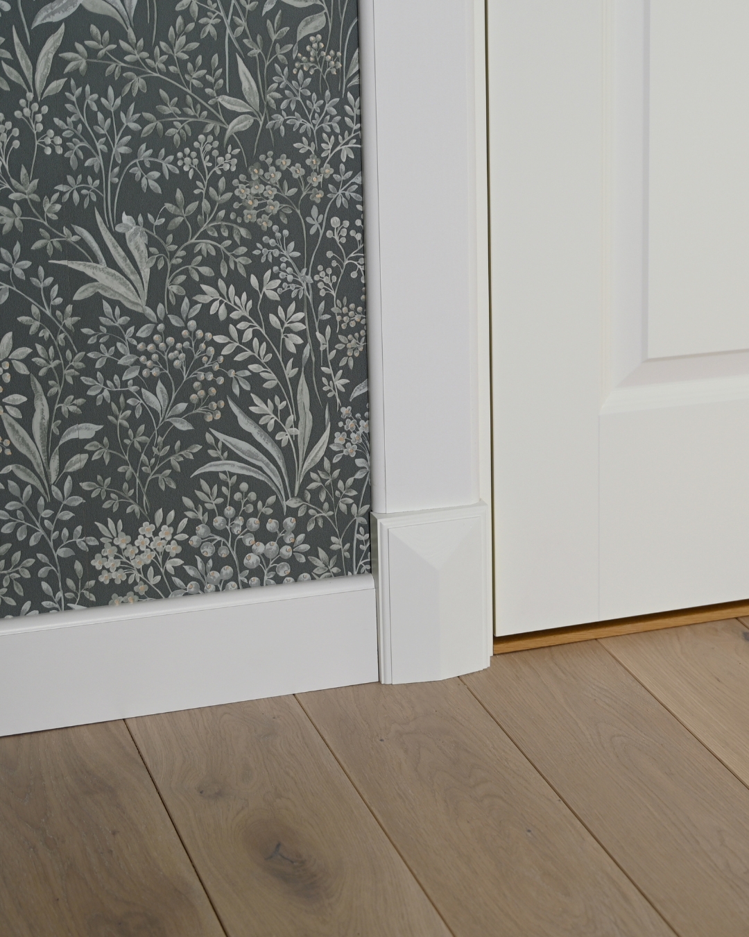 Vägg med vita lister och vit golvkloss, samt en grön tapet med mönster