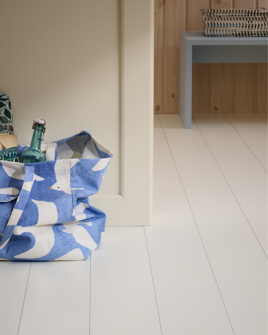 Hörnet av en beige vägg med lister, samt målat trägolv och en blå- och vitrandig väska