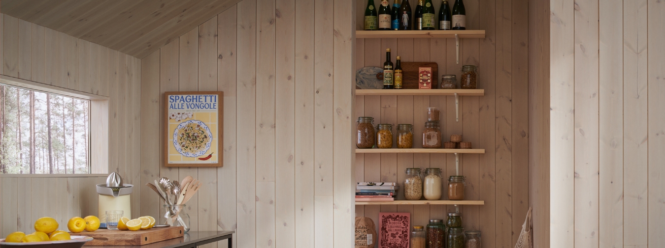 Skafferi med trägolv och ljus träpanel (Stilla Morgondis). Hylla med burkar och flaskor, samt annat för skafferier