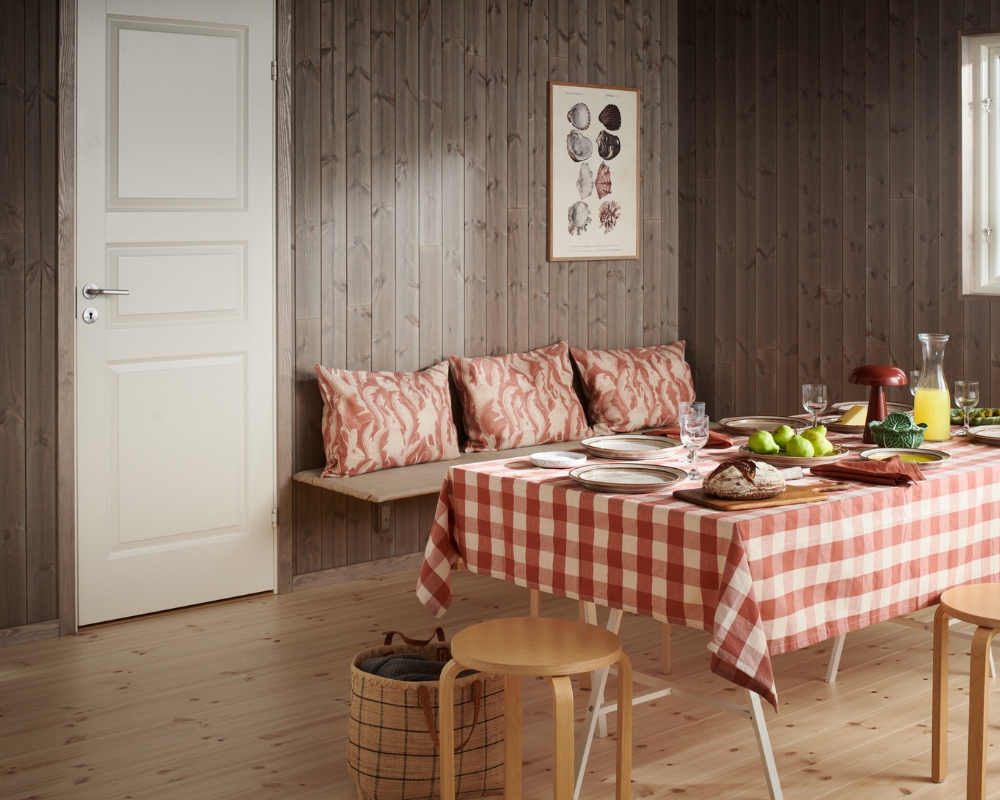 Matbord med vit- och rödrutig duk, samt en bänk på väggen med kuddar. Trägolv och träpanel