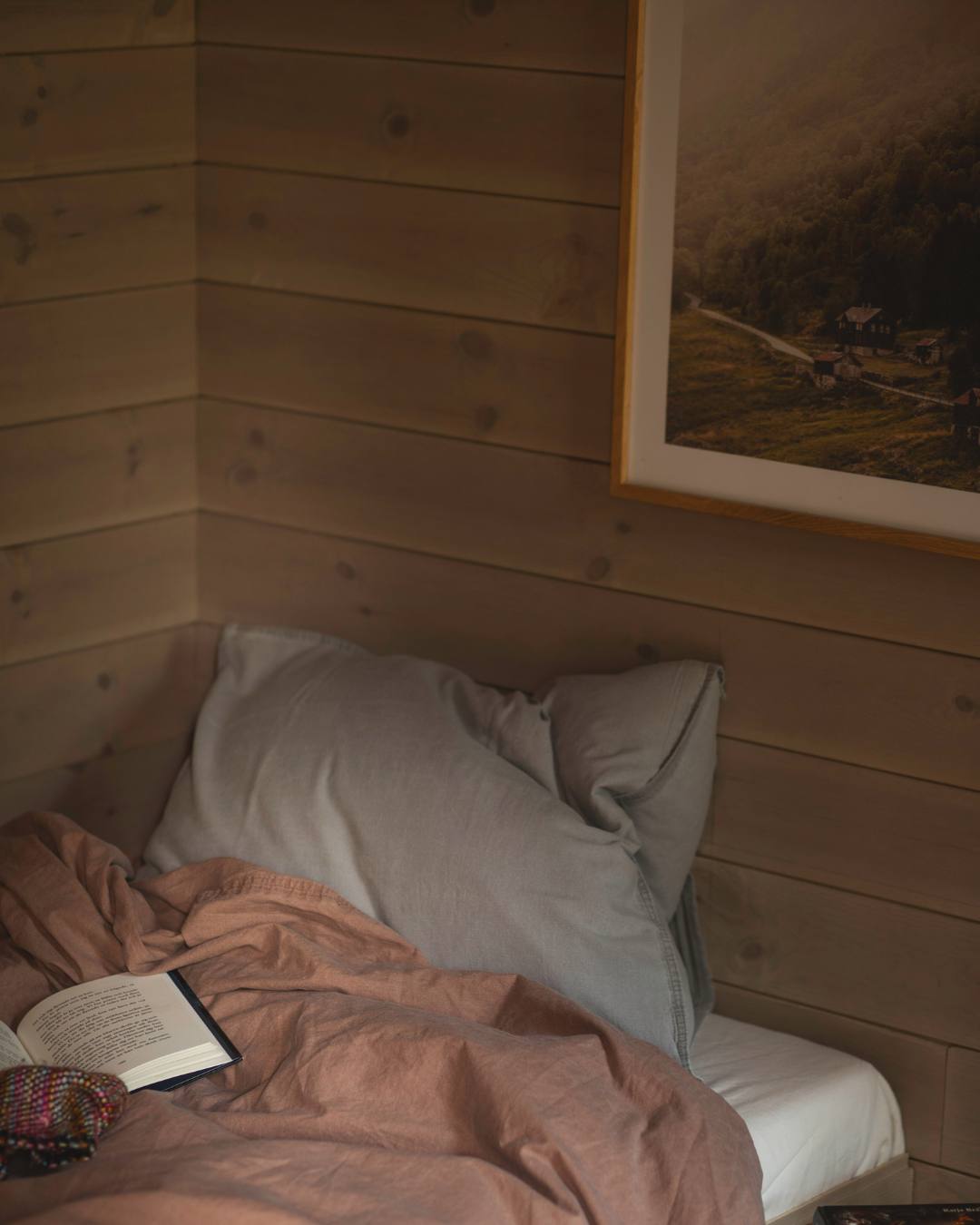 Närbild på ett hörn i ett sovrum med liggande träpanel där panelen möts i linje i hörnet. I den slarvigt bäddade sängen ligger en uppslagen bok.