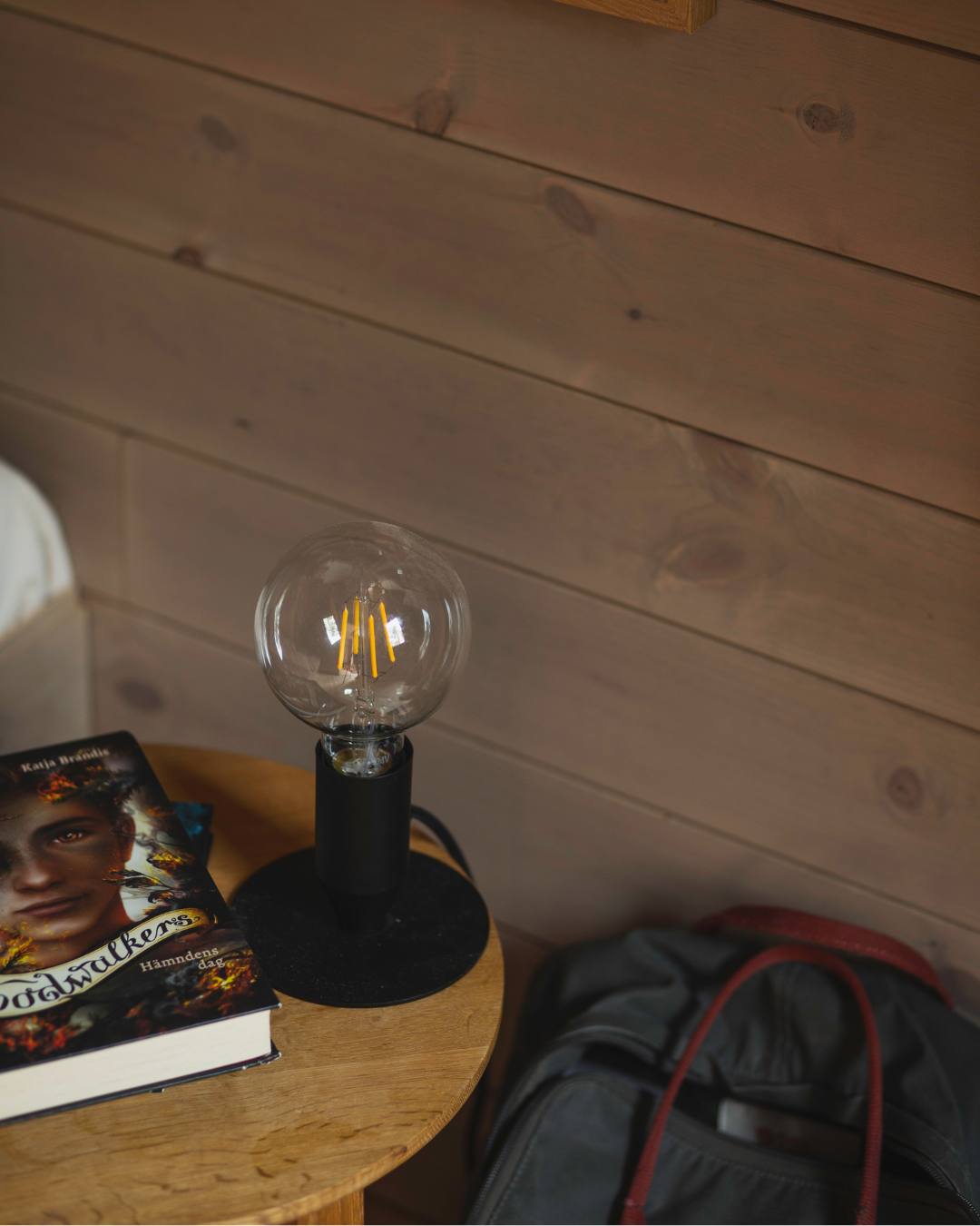 Naken glödlampa i svart fot står på ett runt träbord där det ligger en bok. På väggen liggande träpanel Stilla Sandbeige.
