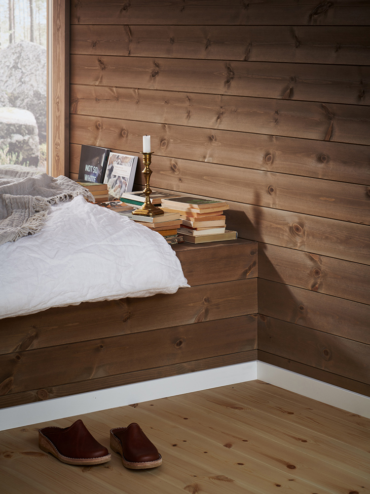 Träpanelen Stilla Skogsbrun sitter på väggarna. Rakt fram ett stort fönster med utsikt mot en tallskog.