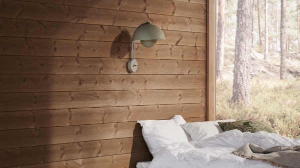 Sovrum med en liggande mörkbrun träpanel på väggen där en sänglampa hänger, i högra hörnet utsikt över en tallskog.