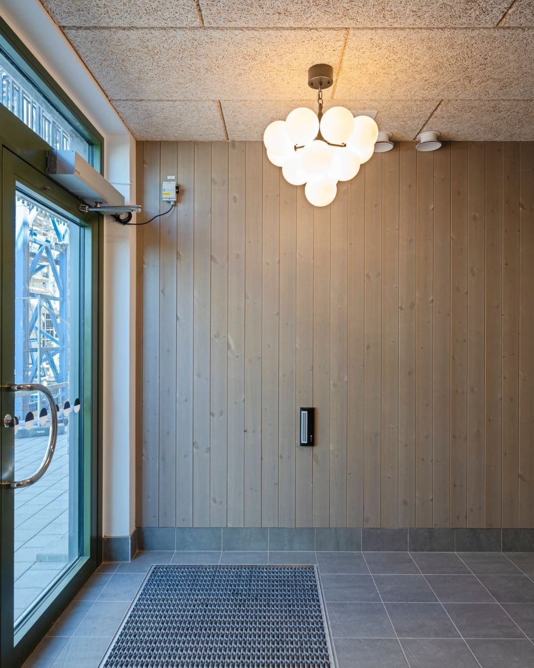 Entren i ett trapphus med dörr till vänster och stående träpanel på väggen raktfram. I taket hänger en lampa.