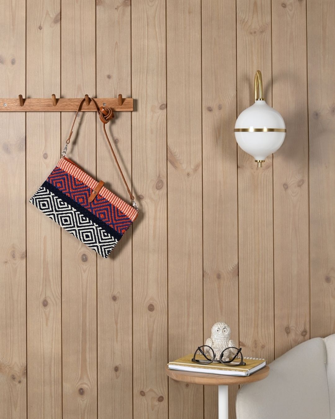 En randig väska hänger på en kroklist av trä bredvid en hängande vi vägglampa. Allt framför en vägg med stående beige panel.