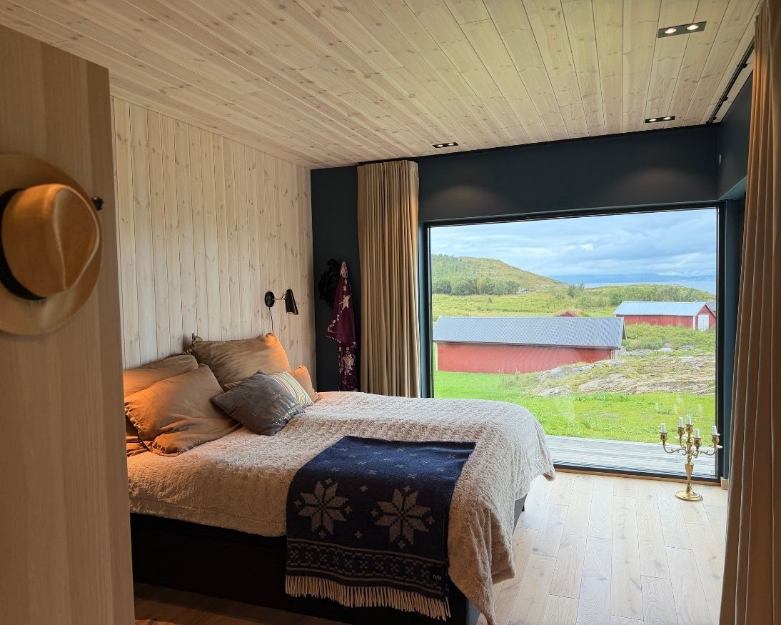 Soverom med lyst trepanel på vegg og i tak, eikegulv og stort vindu med utsikt over landskapet på Rolløya.