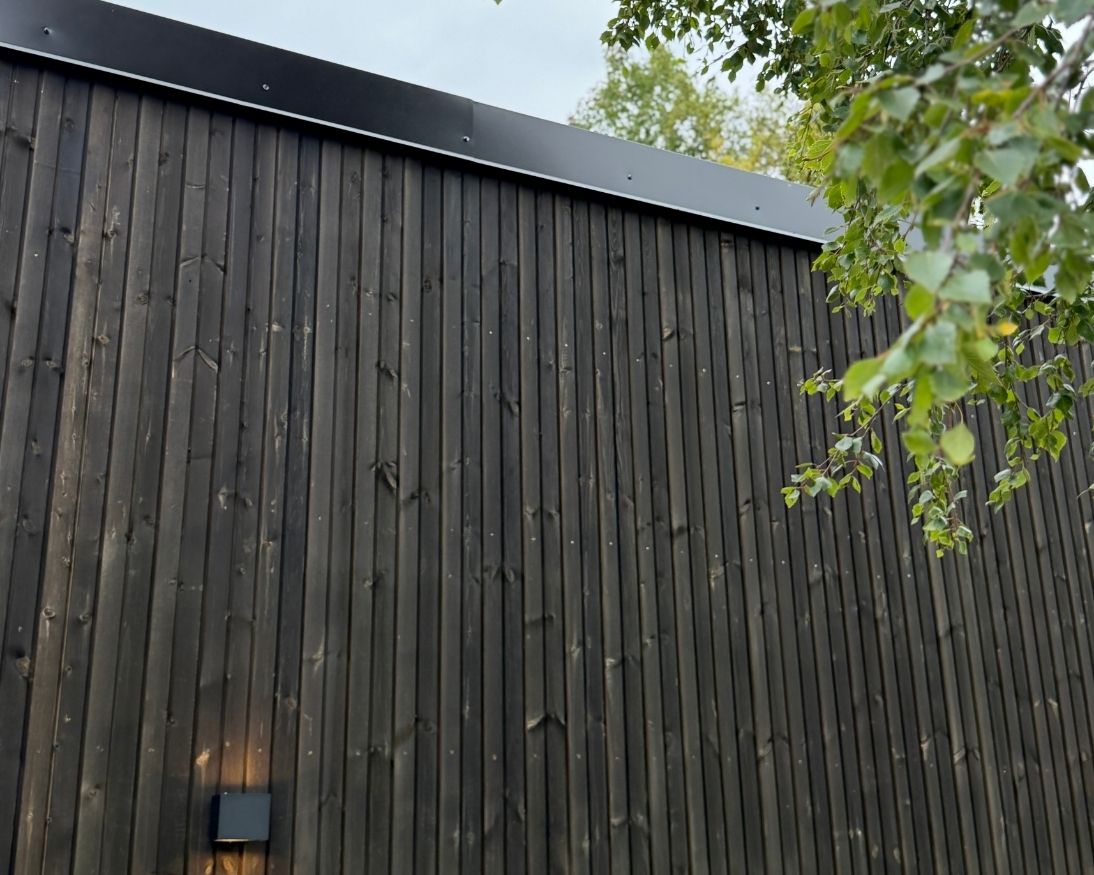 ærbilde av mørk Værbitt-kledning i fargen Garva på fasaden på Haveng, med grønne greiner foran huset.