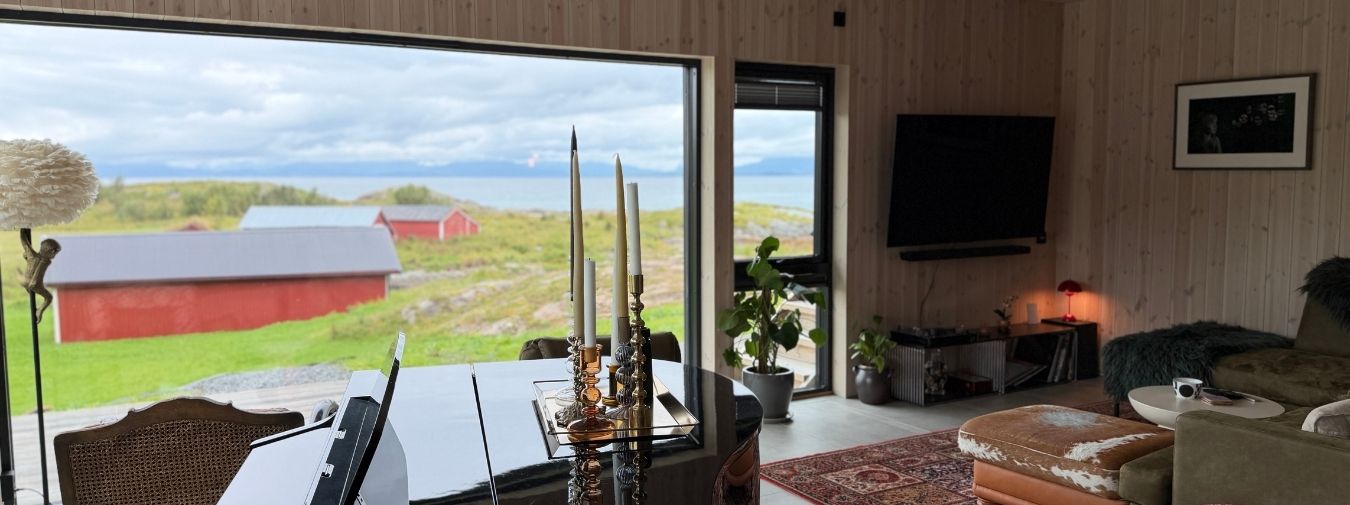 Stue på Haveng med lyst trepanel på vegger og i tak, flygel, sittegruppe og store vindusflater mot naturen på Rolløya.