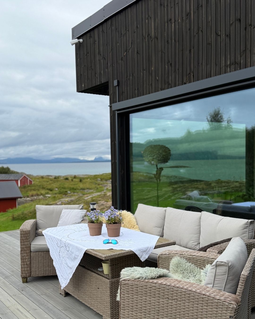 Terrasse med sittegruppe foran den mørke trefasaden på Haveng, med hav og kystlandskap i bakgrunnen.
