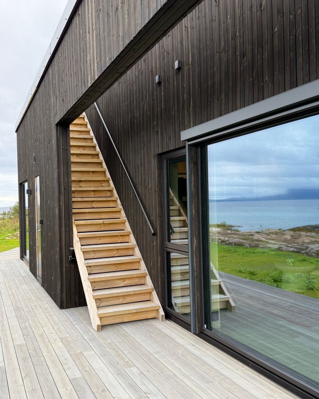 Mørk trekledning, utvendig tretrapp og store vindusflater på Haveng, med terrasse langs huset.