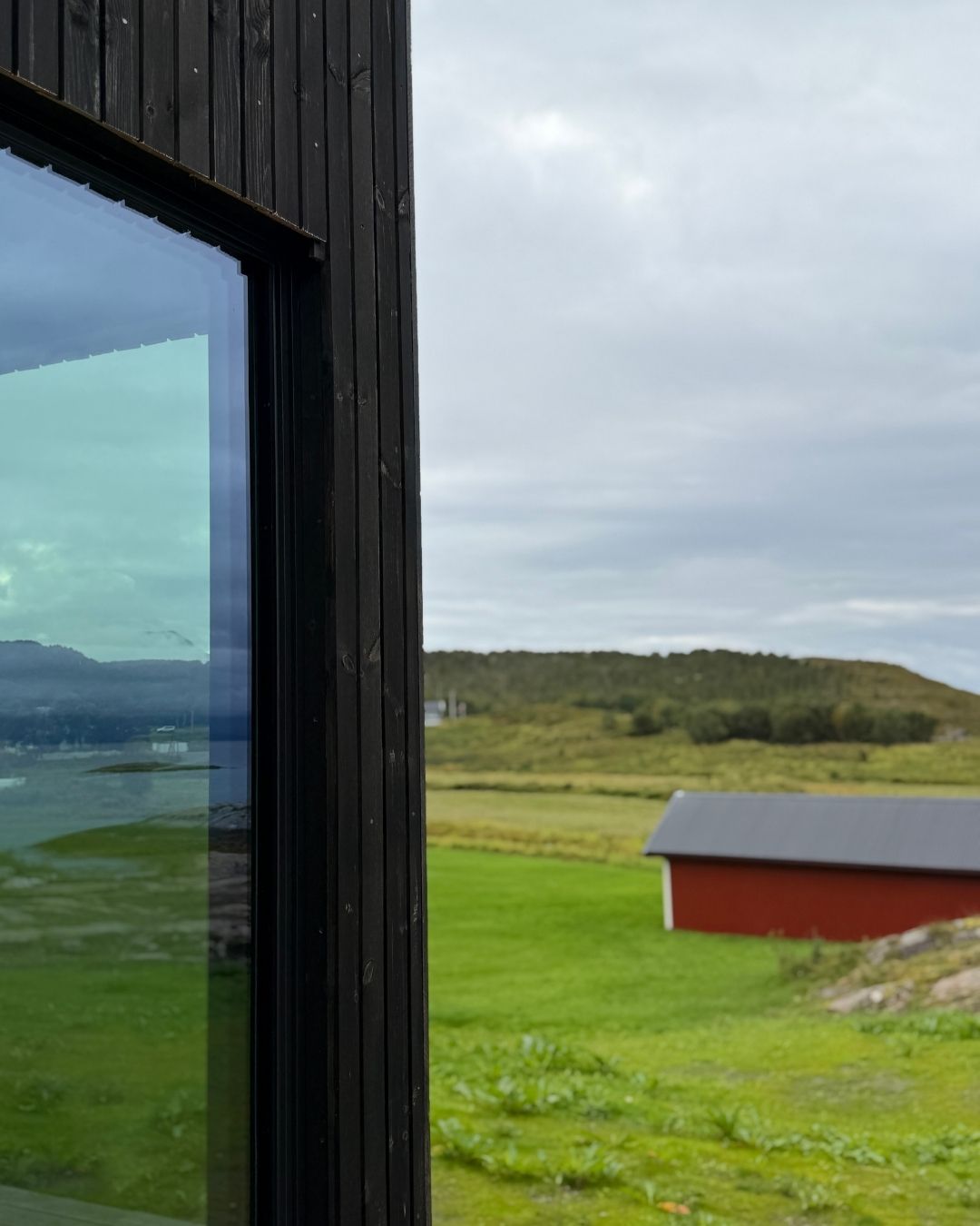 Detalj av mørk trekledning og et stort vindu på Haveng, med grønt landskap og en rød bygning i bakgrunnen.