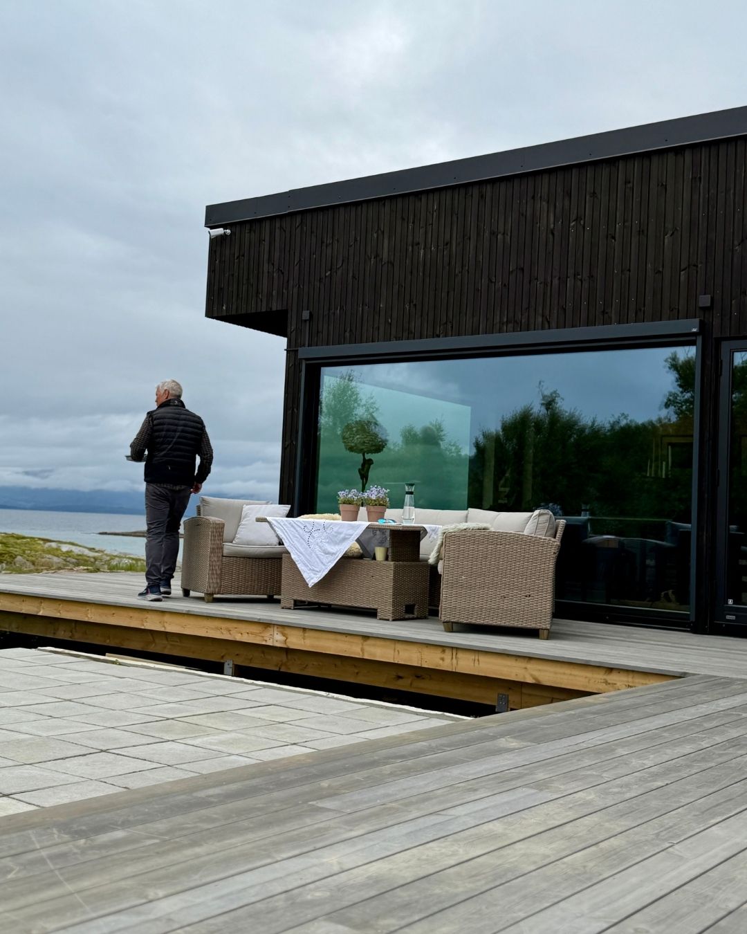 Jan Roger går langs terrassen foran den mørke fasaden på Haveng, med havet i bakgrunnen.