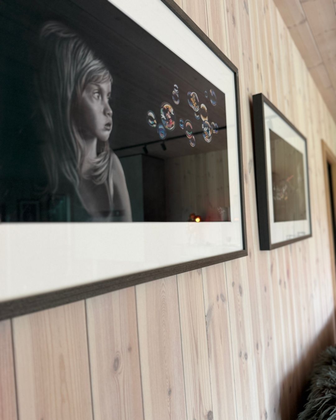 Innrammede kunstbilder henger på en vegg med lyst trepanel.