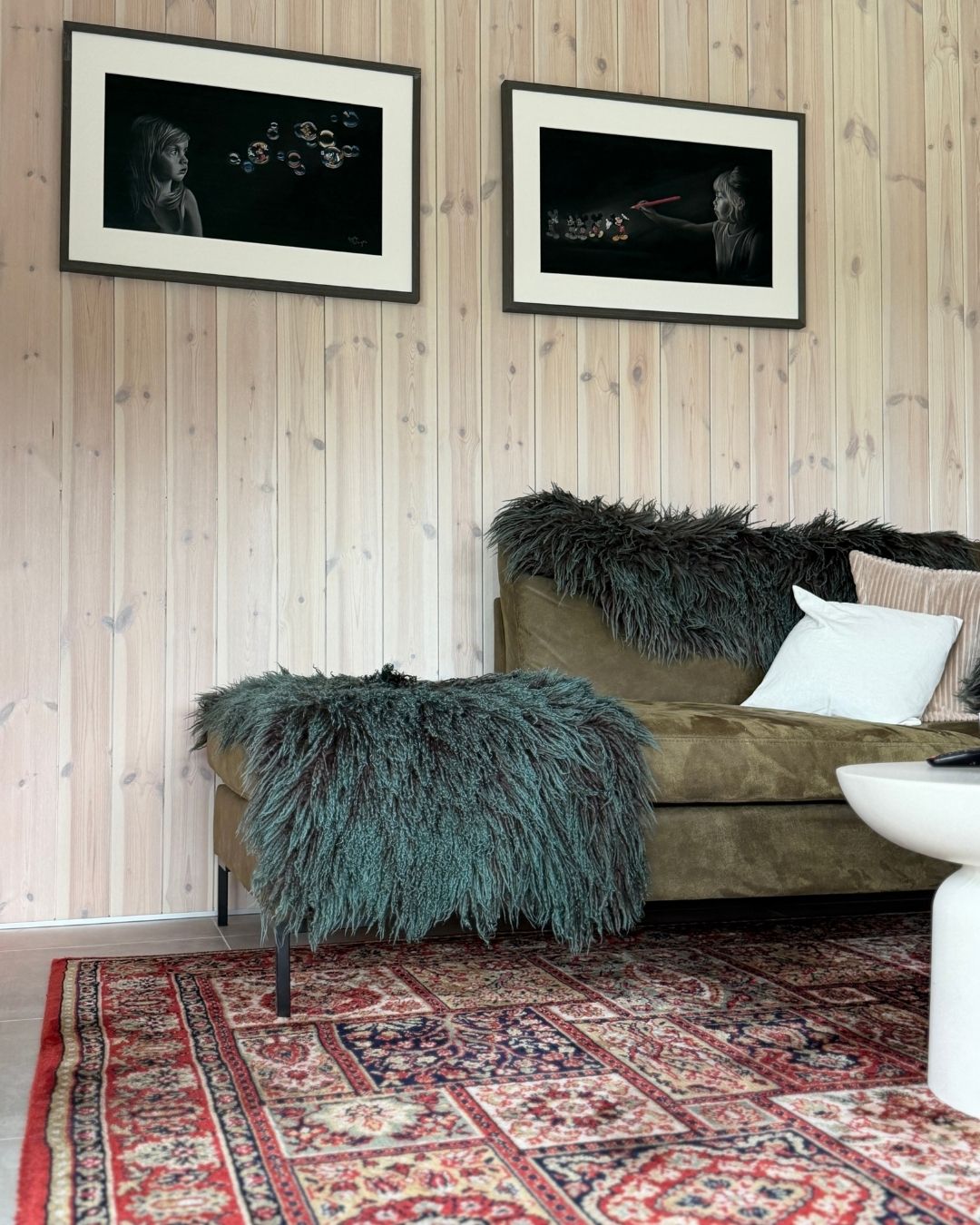 Grønn sofa med puter og pledd foran en vegg med lyst trepanel og innrammede bilder.