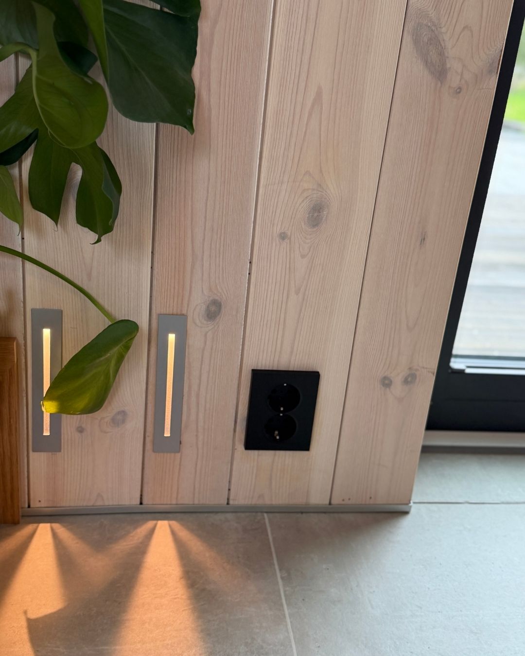Nærbilde av lyst trepanel med stikkontakt, innfelte lys og grønne planteblader.