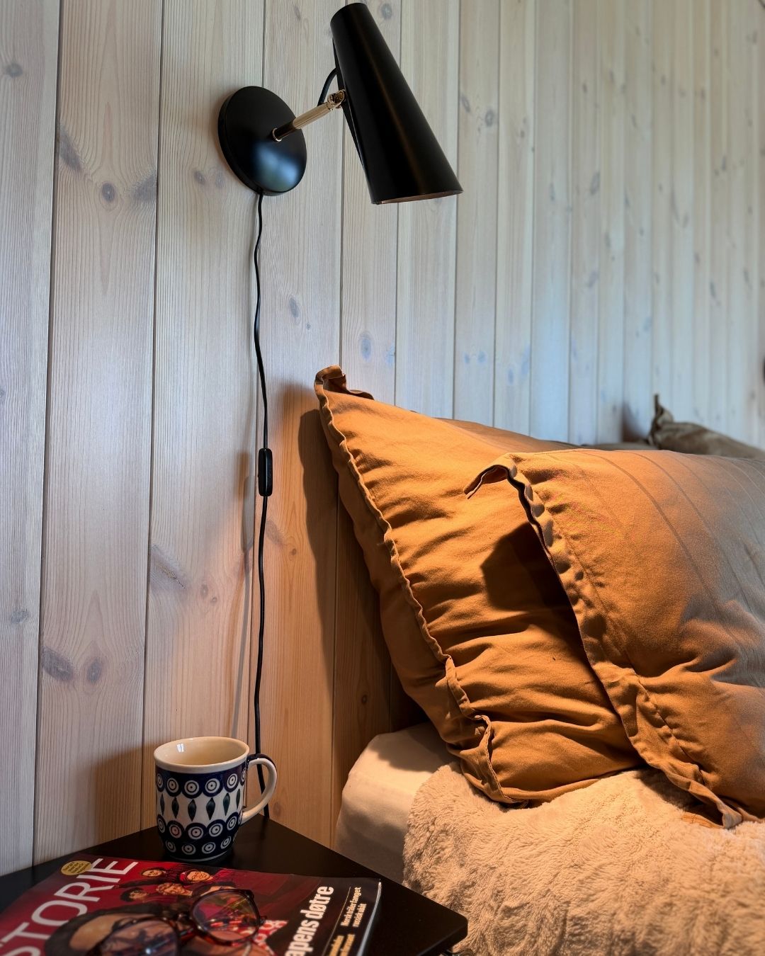 Seng med varme tekstiler og svart vegglampe montert på lyst trepanel.