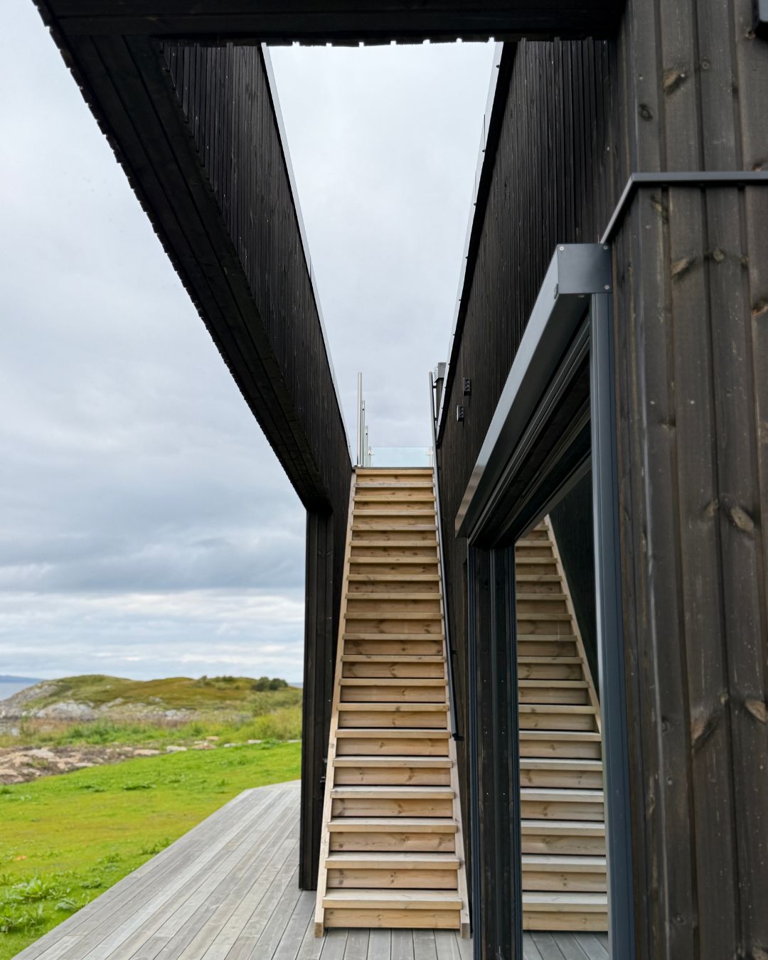 Tretrapp mellom to mørke bygningsvolumer på Haveng, omgitt av terrasse og kystlandskap.