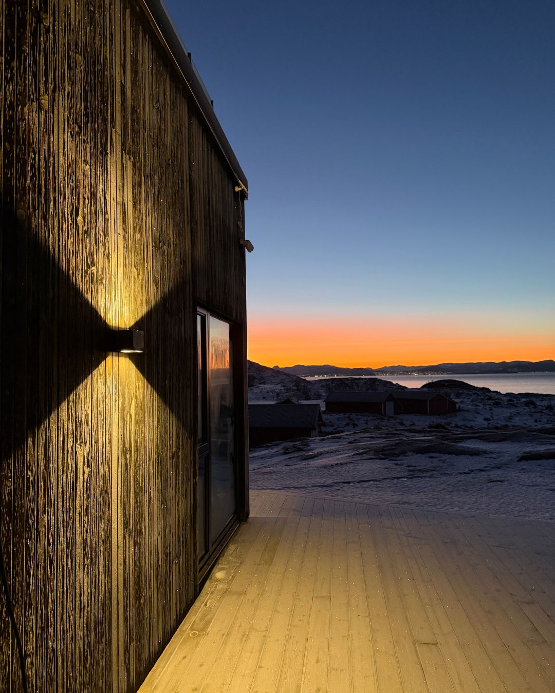 Mørk trefasade på Haveng i vinterlandskap, med solnedgang over havet i bakgrunnen.