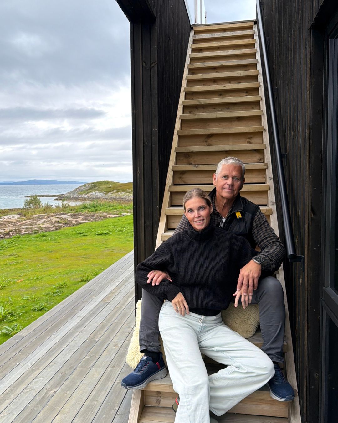 Hege og Jan Roger sitter sammen i trappen ved Haveng, med terrasse, hav og kystlandskap rundt seg.