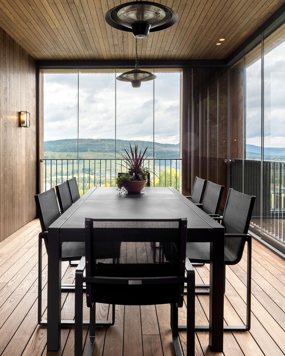Overdekket terrasse med tak og vegger i Moelven termoask, stor svart spisestue-gruppe for åtte og panoramautsikt mot åslandskap gjennom glassfelt og rekkverk.