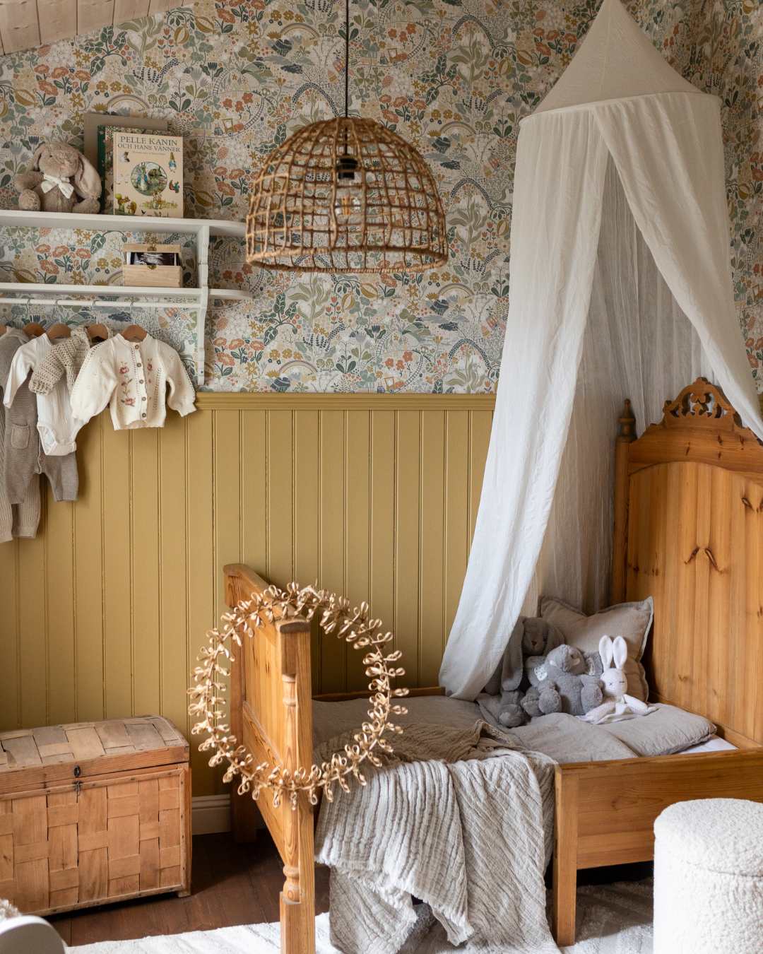 Barnerom med himmelseng og leker- Veggen har høy brystingspanel i en gul stående trepanel, siste del av veggen har en blomstet tapet. Lampe i naturmateriale.