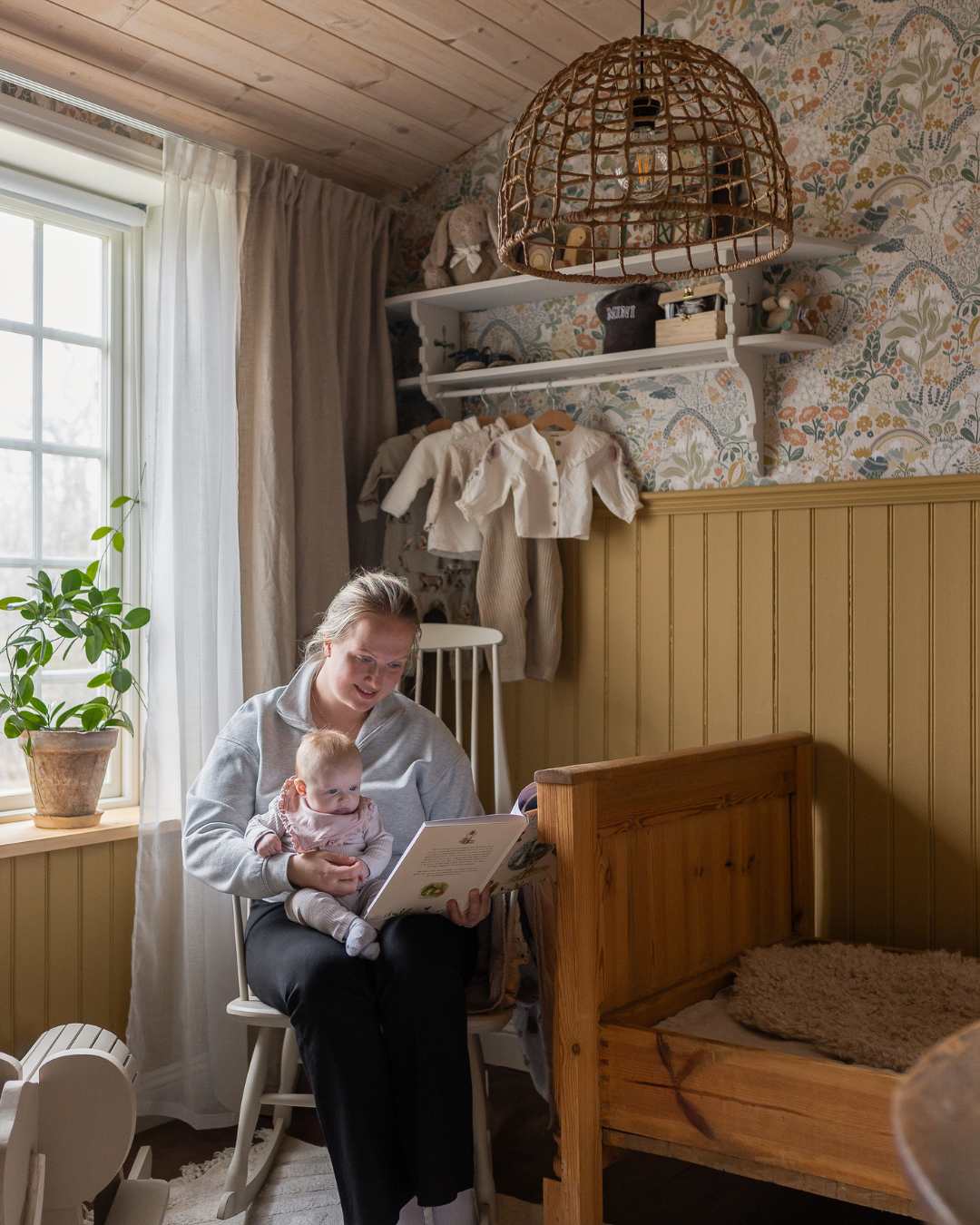 Kvinne med baby på fanget i et barnerom med hvitlistet vindu og gul stående brystningspanel