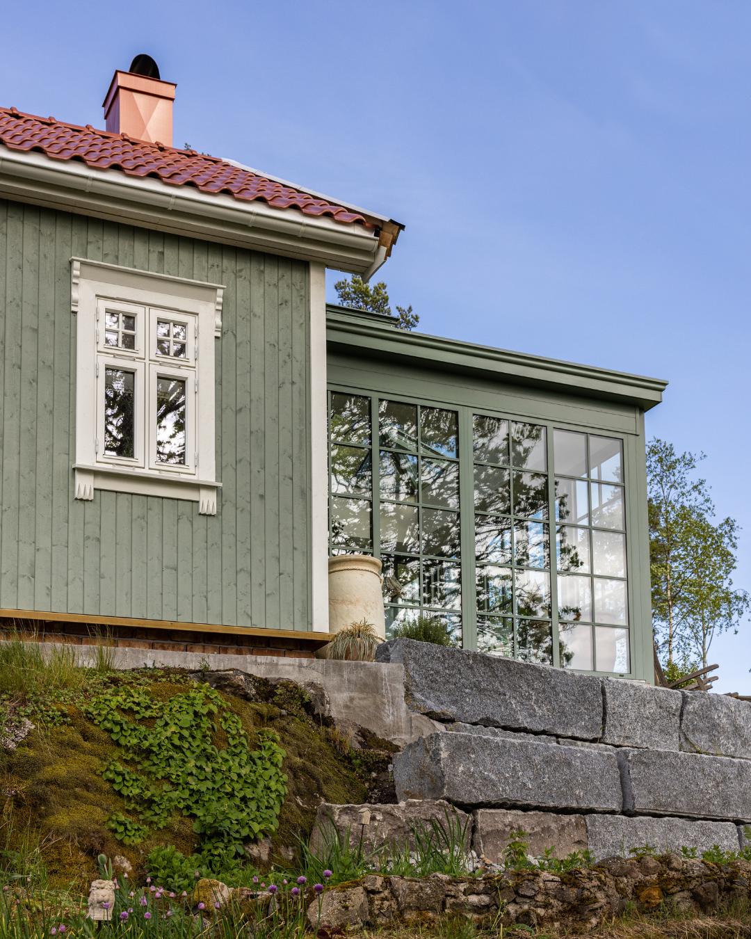 Fasade på grønnmalt trehus med tegltak og orangeri med store sprossevinduer, sett fra natursteinsmur med blomstrende hage i forgrunnen.