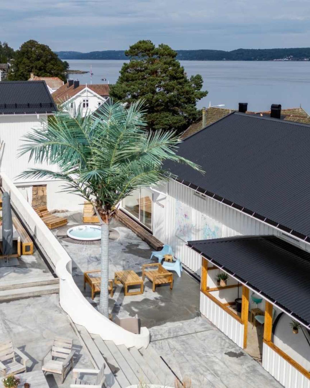 Fugleperspektiv over Villa Oslofjord med palmer, boblebad, utemøbler og Oslofjorden i bakgrunnen. Bygningen har hvit kledning og svart tak.