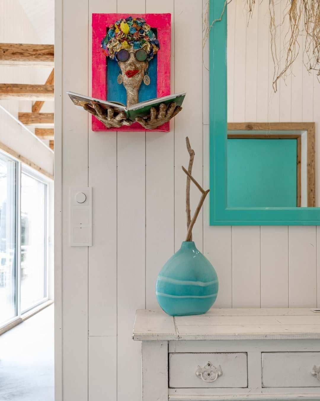Inngangsparti i Villa Oslofjord med hvit Sprekkpanel, en turkis vase på en gammel kommode, turkisfarget speilramme og et fargerikt collage-kunstverk på veggen.