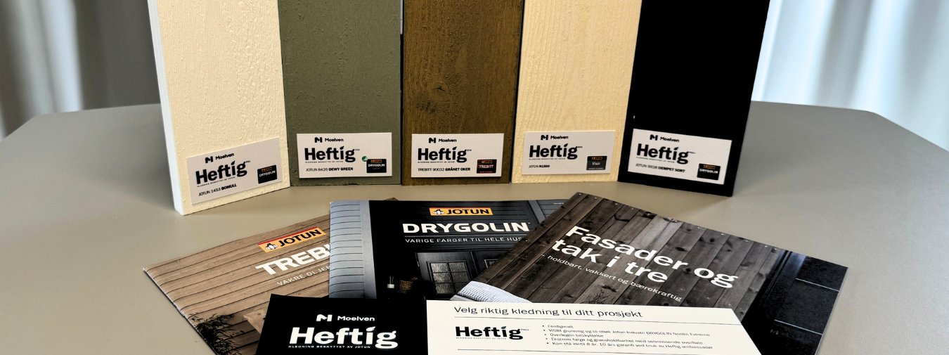 Fem prøvebiter av Heftig kledning i ulike farger og overflater stilt opp på et bord, foran ligger brosjyrer og fargehjelpemidler fra Moelven og Jotun – inkludert DRYGOLIN fargekart, TREBITT fargekart, fasadekatalog og et veiledningsark for valg av Heftig kledning.
