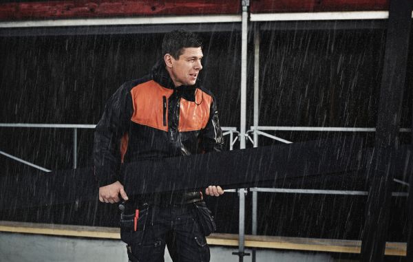 En mann iført arbeidsjakke i sort og oransje håndterer ytterpanel utendørs i kraftig regnvær. Han holder i en planke og arbeider på et bygg- eller anleggssted, med mørk bakgrunn og synlige stålkonstruksjoner.
