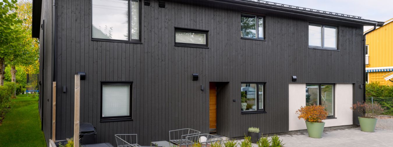Enebolig i to etasjer sett fra hagesiden. Fasaden er kledd med Heftig Pro i fargen Labrador, kombinert med hvit murpusset veggflate og svarte vinduer. Naturtre inngangsdør, utemøbler på terrassen og potteplanter ved inngangen. Grønn hage og nabobygg synlig i bakgrunnen.