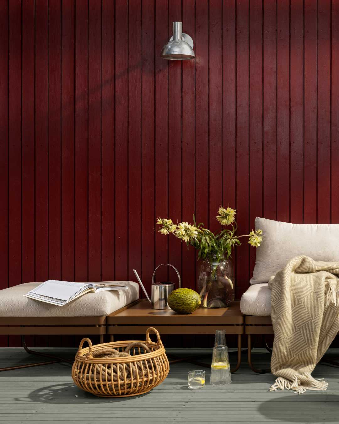 Kledningsvegg i tre med stående kledning i fargen mørk rød, benk med puter og tepper forann.