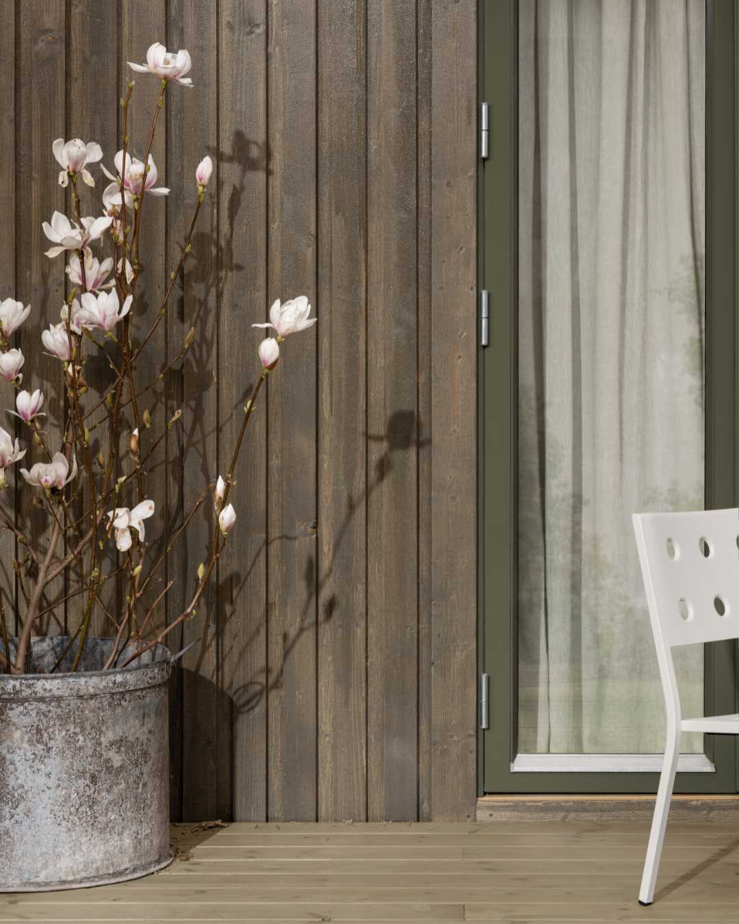 Fasade med ståemde gråbeiset trekledning. Terrasse stol og blomstrende kirsebærtre i forkant