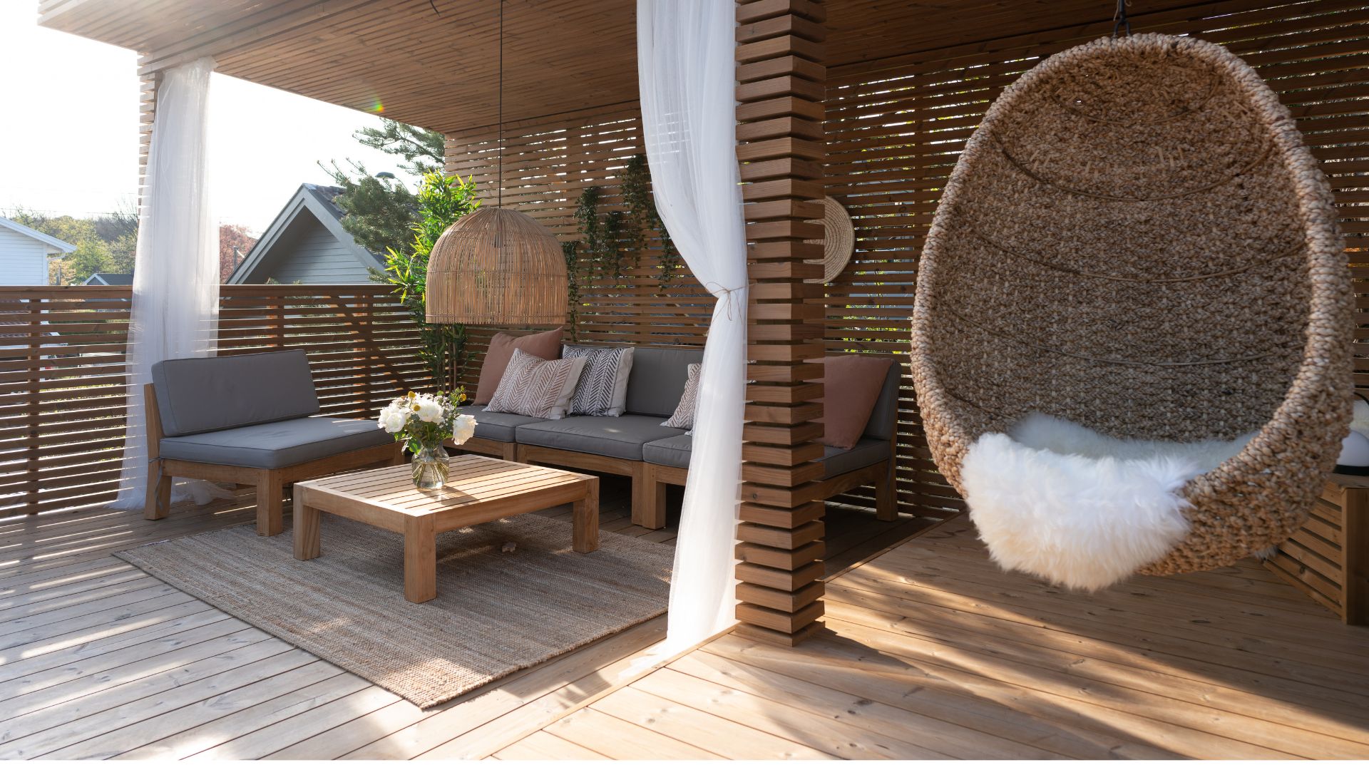 Detaljert utsnitt av sittegruppe på termoterrasse fra Moelven med trebenk, puter, lavt bord, hvite gardiner og en flettet hengestol.