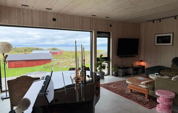 Stue på Haveng med lyst trepanel på vegger og i tak, flygel, sittegruppe og store vindusflater mot naturen på Rolløya.