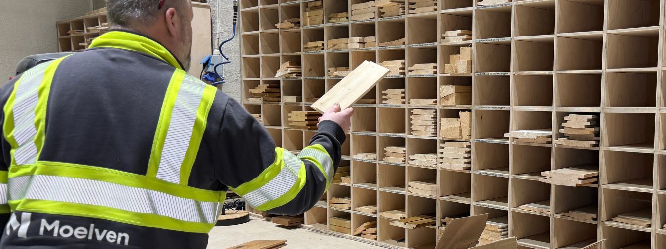 En Moelven-ansatt i gul refleksvest holder opp en treprøve foran et stort reolanlegg fylt med ulike treprøver og trefargepaneler, organisert i mange små hyller.