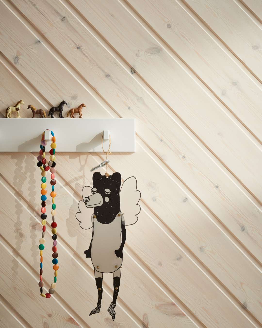 Trepanel Skygge skrå i hvitlasert montert diagonalt, med knaggrekke der det henger et smykke og en papirfigur. Oppå knaggen står det plastleker av hester.